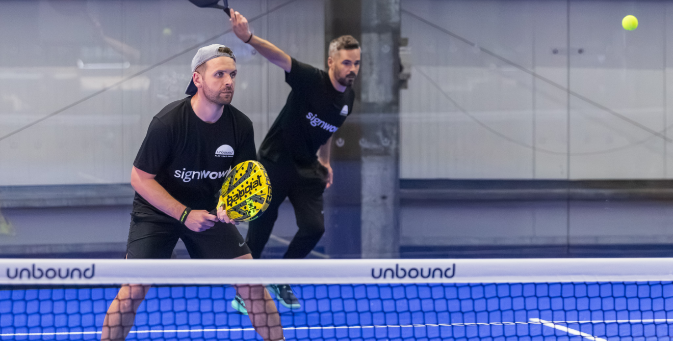 Mae dau chwaraewr padel byddar yn sefyll y tu ôl i'r rhwyd. Mae un yn cymryd ergyd tra bod y llall yn aros.