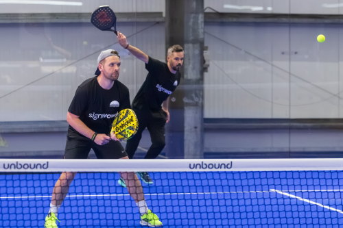 Mae dau chwaraewr padel byddar yn sefyll y tu ôl i'r rhwyd. Mae un yn cymryd ergyd tra bod y llall yn aros.